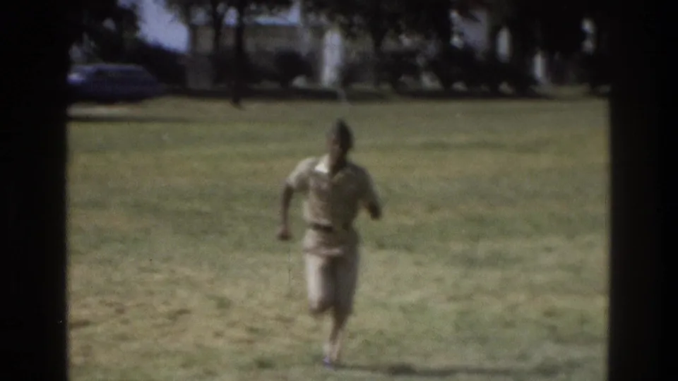 Black uniformed man running toward the camera across a grassy Atlanta field in faded 1975 8mm footage.