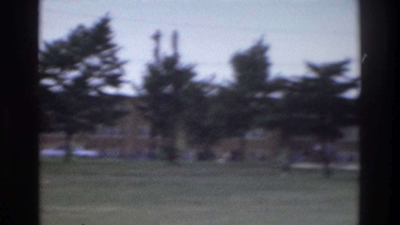 1975 Atlanta home-movie frame showing a grassy roadside area with trees, utility poles, and a low brick building seen through dark vertical edges.