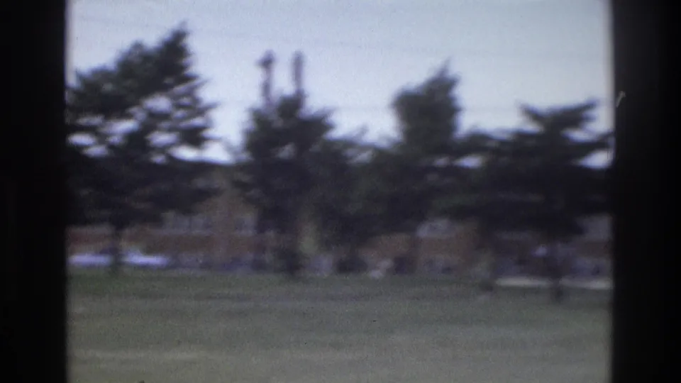 1975 Atlanta home-movie frame showing a grassy roadside area with trees, utility poles, and a low brick building seen through dark vertical edges.