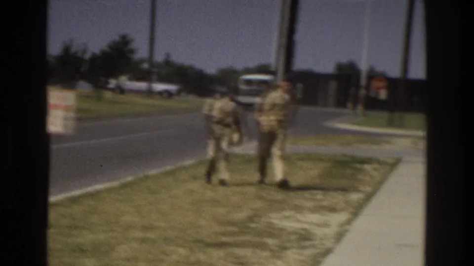1975 Atlanta home-movie frame showing two people in light clothing on a sidewalk beside a road, with utility poles and a distant bus-like vehicle nearby.