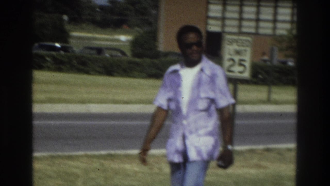 In 1975 Atlanta home-movie footage, a man in sunglasses and a light shirt walks toward the camera near a road and speed-limit sign.