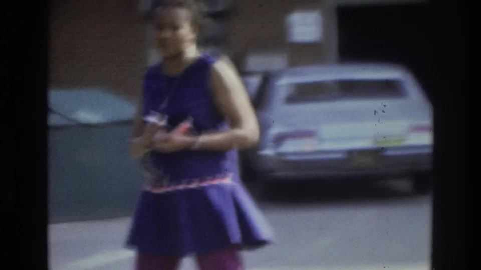 In 1975 Atlanta footage, a woman in a purple sleeveless dress walks outdoors with parked cars and brick buildings behind her.