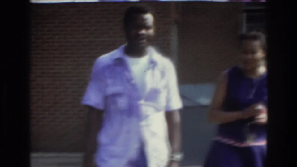 The man in a light shirt and the woman in a purple dress pass near a brick building in Atlanta home-movie footage from 1975.