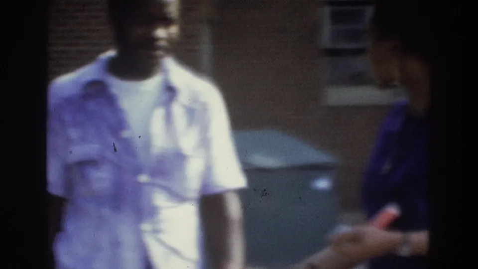A man in a light short-sleeve shirt is framed beside a woman partly visible at the edge, with brick building and parked car behind.