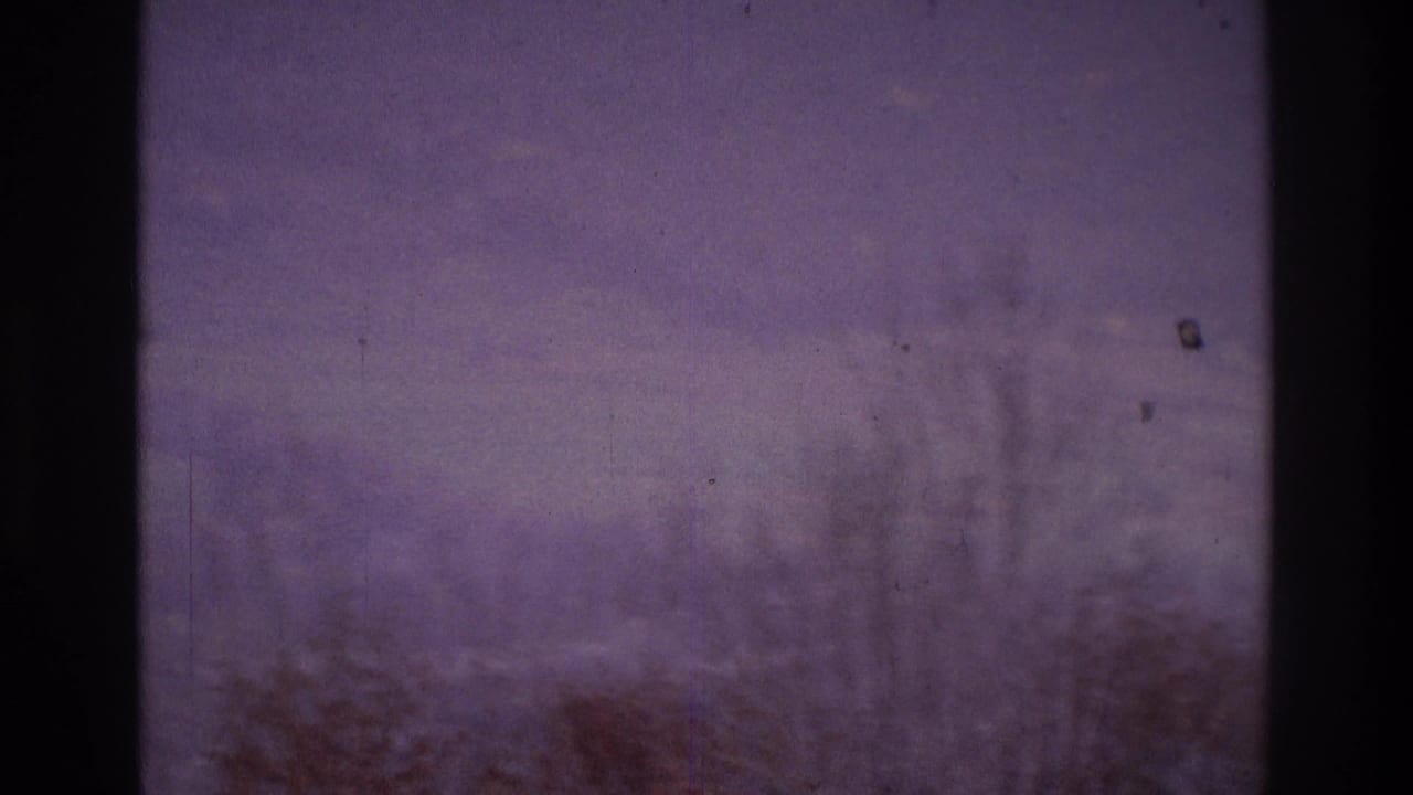 1975 Atlanta-area 8mm frame looking through a window at bare trees, haze, and a faint hillside settlement in the distance.