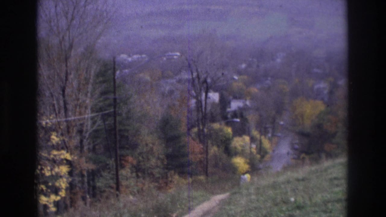 1975 Atlanta-area 8mm frame looking down a grassy slope toward a dirt path, utility poles, autumn trees, and distant houses.