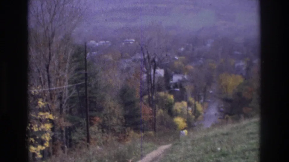 1975 Atlanta-area 8mm frame looking down a grassy slope toward a dirt path, utility poles, autumn trees, and distant houses.