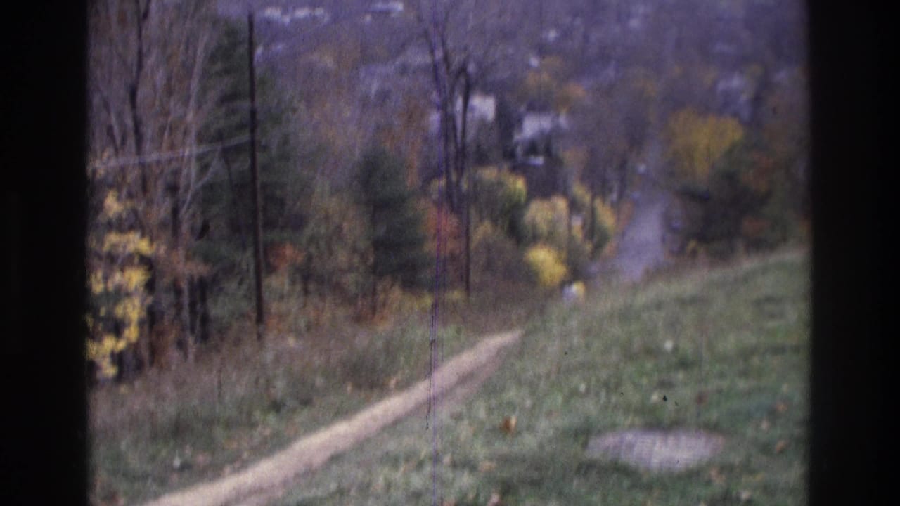 1975 Atlanta home-movie frame showing a dirt path, autumn trees, and distant residential buildings beyond a grassy slope.