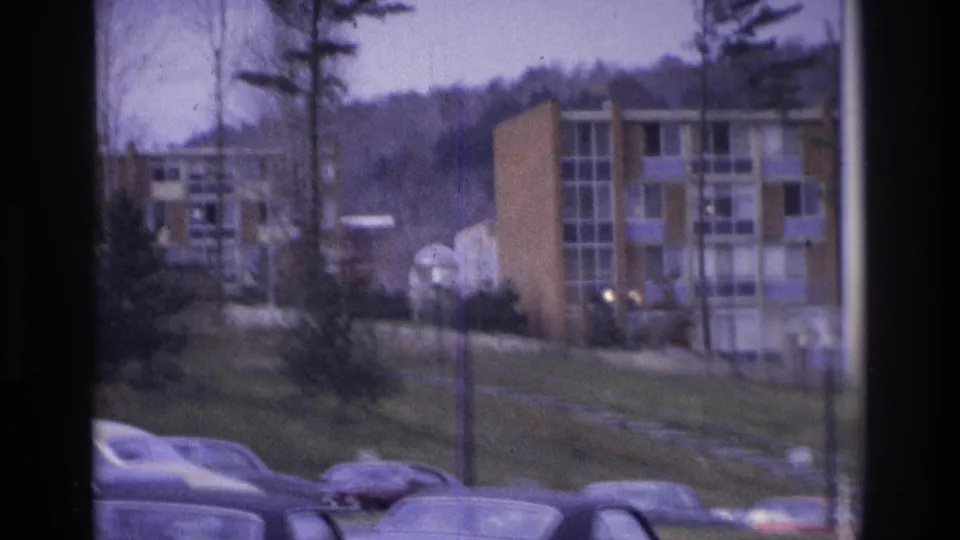 1975 Atlanta home-movie frame with apartment blocks, parked cars, lampposts, and a wooded hillside in the background.