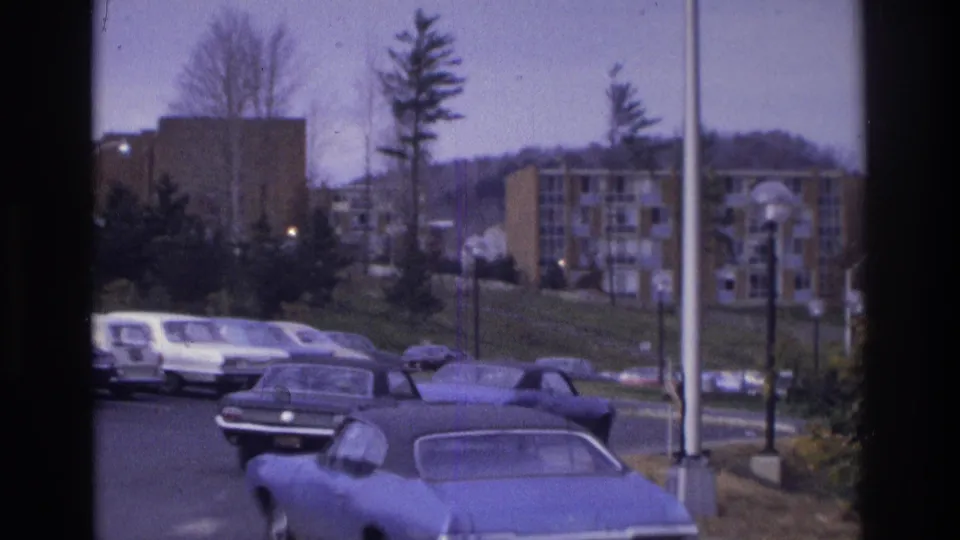 1975 Atlanta residential scene with parked cars, a white van, trees, lampposts, and brick apartment buildings in soft 8mm color.