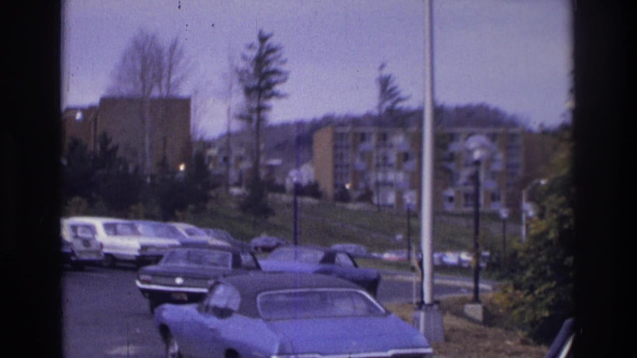 1975 Atlanta home-movie frame showing a suburban apartment parking lot with period cars, trees, light poles, and buildings on a rise.