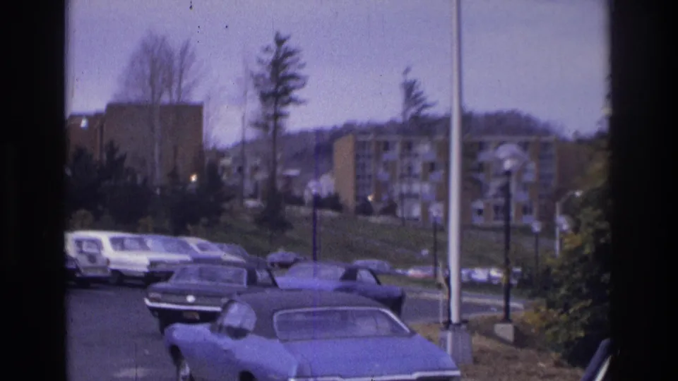 1975 Atlanta home-movie frame showing a suburban apartment parking lot with period cars, trees, light poles, and buildings on a rise.