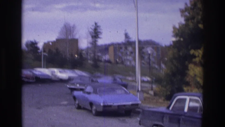 1975 Atlanta home-movie view across a suburban parking lot, with a blue car in front and apartment buildings visible through soft focus.