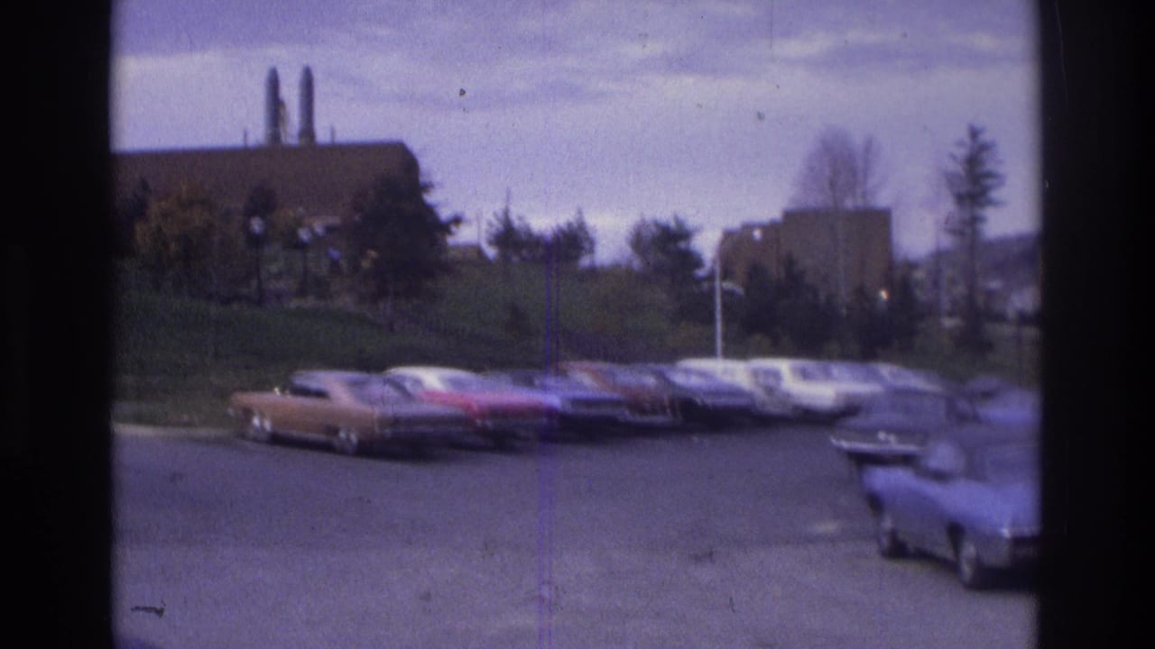 Atlanta 1975 home-movie frame showing classic cars parked on asphalt near a grassy slope and brick building with smokestacks.