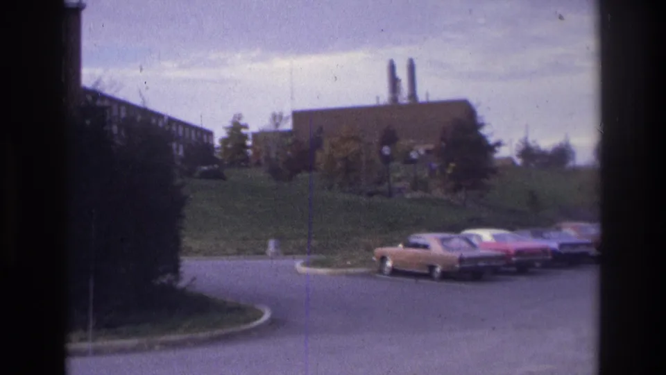 Home-movie frame from Atlanta in 1975 showing a curved drive, parked sedans, grassy landscaping, and a brick building with stacks.