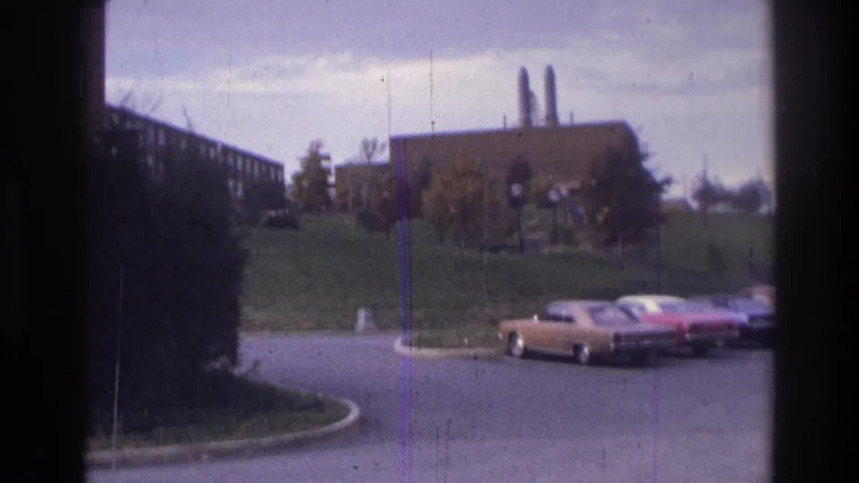 Atlanta 1975 8mm frame with parked cars in the foreground and a brick building with twin smokestacks beyond a grass bank.