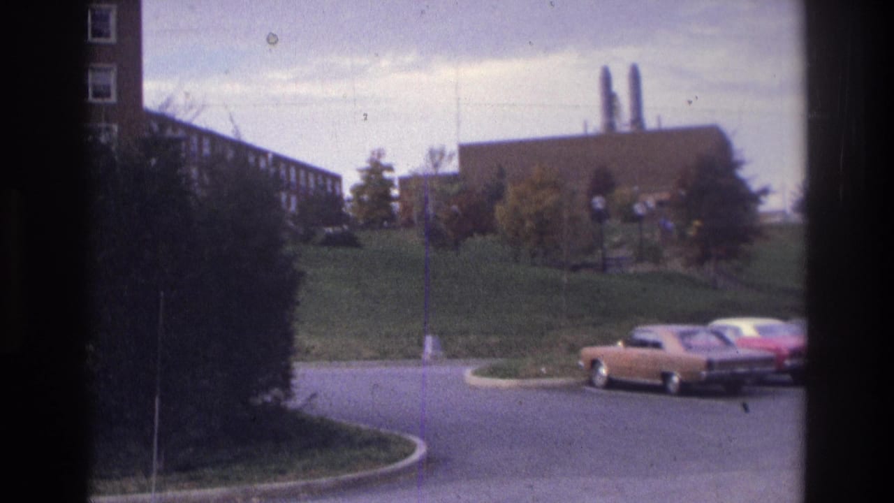 1975 Atlanta home-movie frame showing a parked sedan near a curving road, brick buildings, green lawn, shrub, and smokestacks beyond.