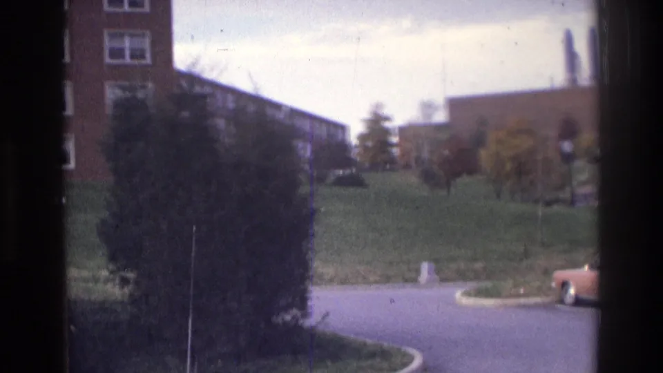 1975 Atlanta 8mm frame with a road curve, large evergreen shrub, grassy slope, brick building edge, and partial parked car.
