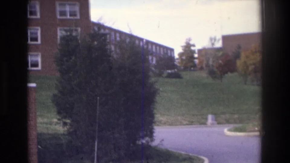 1975 Atlanta home-movie frame looking past a large shrub and road curve toward a brick apartment building and grassy slope.