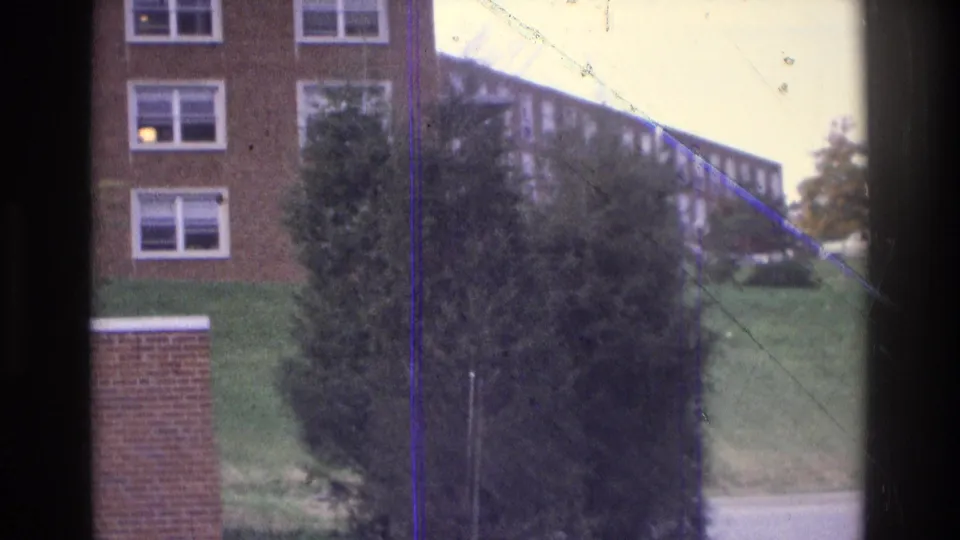 1975 Atlanta 8mm frame showing brick building windows, a large shrub, sloped lawn, low wall, road edge, and distant institutional building.