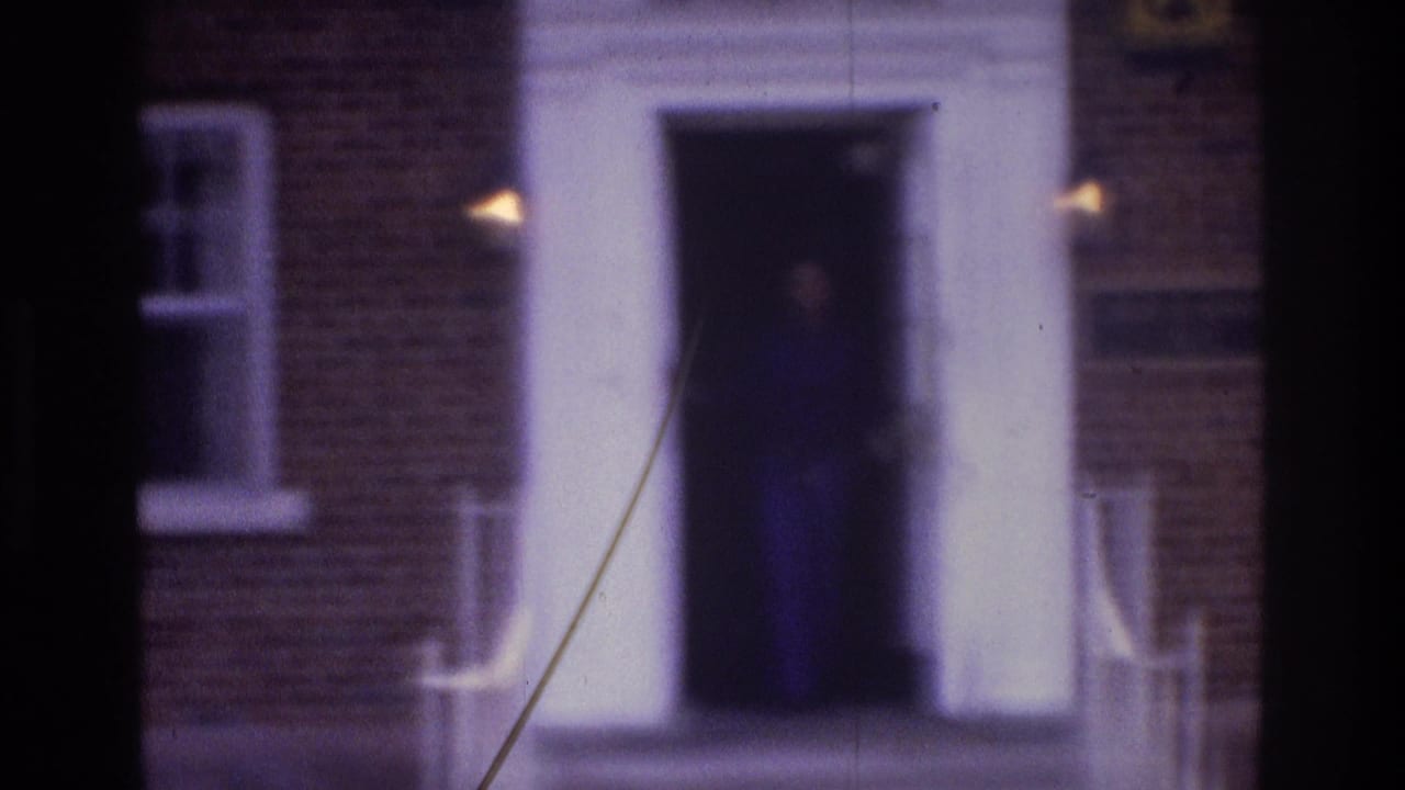 Soft 8mm frame of a white apartment doorway on a brick building, with a woman barely visible in the dark entrance.