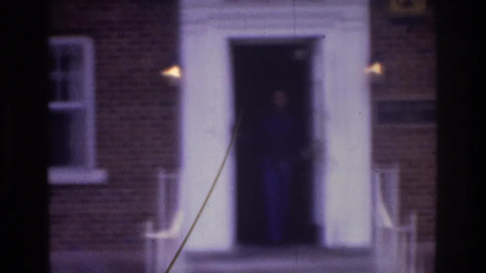 Soft 8mm frame of a white apartment doorway on a brick building, with a woman barely visible in the dark entrance.