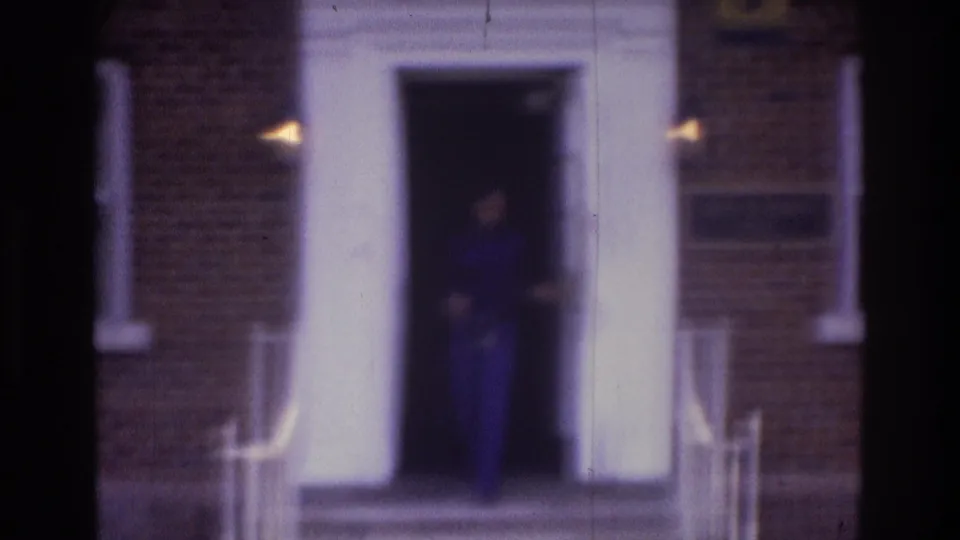 Atlanta 1975 home-movie frame showing a woman in blue clothing at a white doorway on a brick apartment building.
