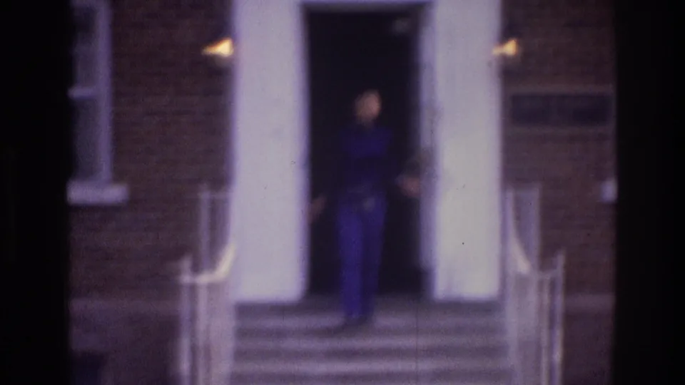 Atlanta 1975 8mm frame of a woman in a blue sweater and jeans descending steps from a white brick-apartment doorway.