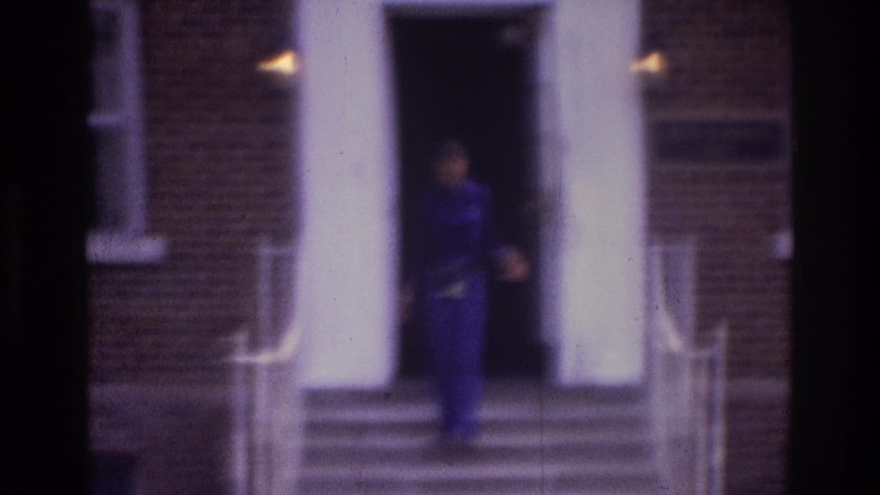 1975 Atlanta home-movie frame showing a person in dark blue near a brick building entrance with white columns and exterior steps.