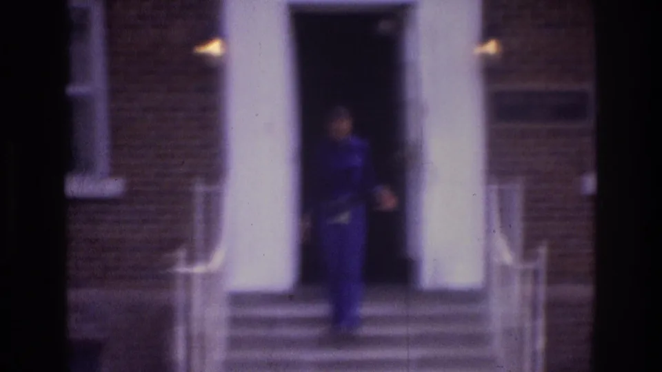 1975 Atlanta home-movie frame showing a person in dark blue near a brick building entrance with white columns and exterior steps.