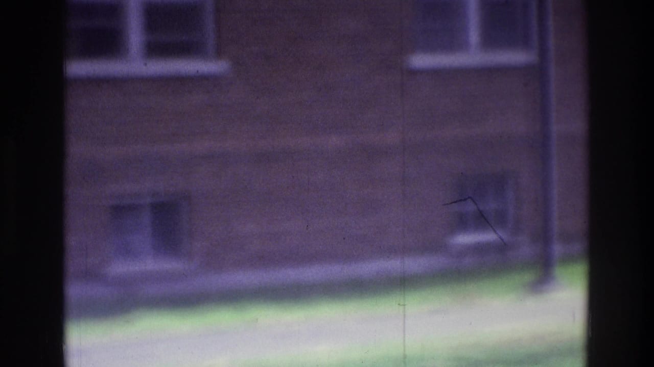 1975 Atlanta 8mm frame showing a soft, distant courtyard view with brick walls, white windows, grass, and a dark vertical film edge.