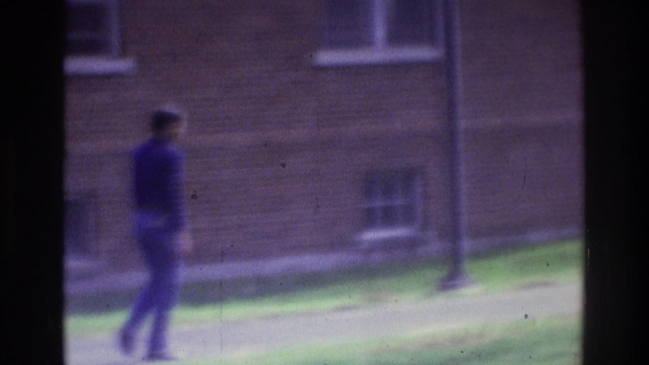 Blurred person in dark clothing walking past a brick building and lampposts.