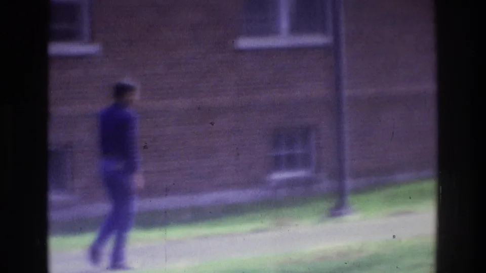 1975 Atlanta 8mm frame with an unidentifiable person walking across a grassy courtyard before a brick building.
