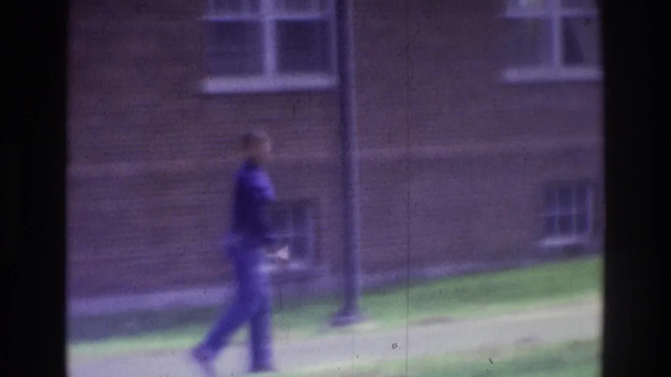 1975 Atlanta 8mm frame showing a distant, unidentifiable person walking past grass and windows in a brick courtyard setting.