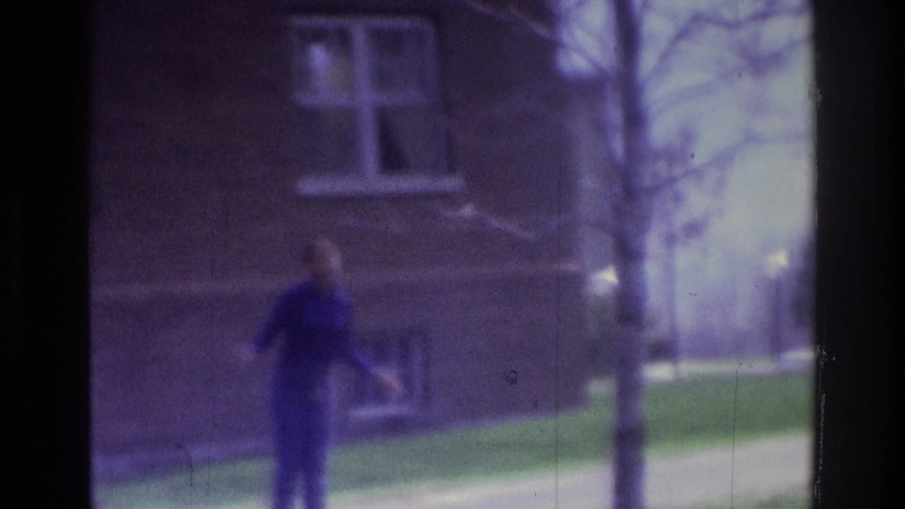 1975 Atlanta 8mm frame showing a person walking past a brick building exterior with windows, grass, and a sidewalk.