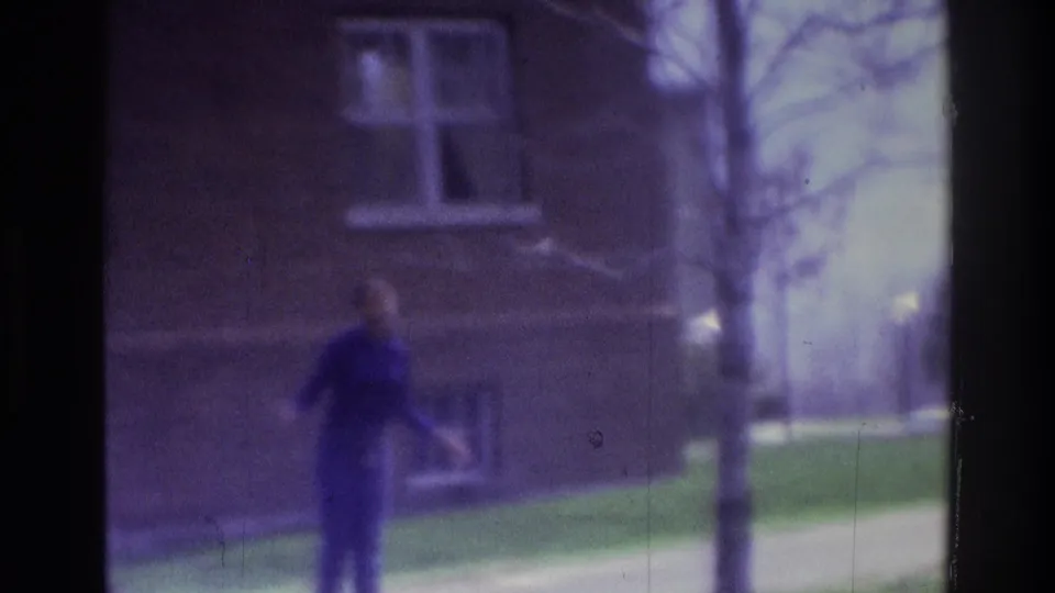 1975 Atlanta 8mm frame showing a person walking past a brick building exterior with windows, grass, and a sidewalk.