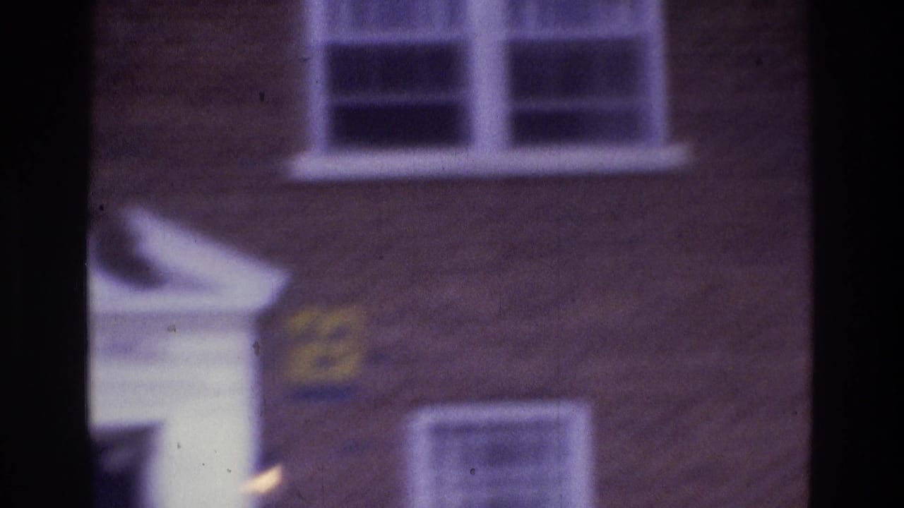 1975 Atlanta home-movie frame showing a brick exterior, white-trimmed windows, and a yellow radiation warning sign near a portico edge.