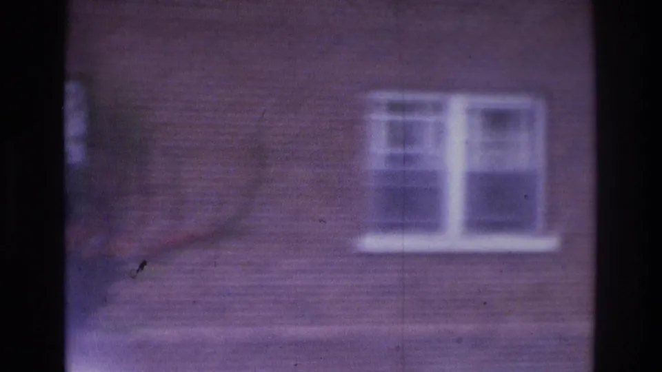 1975 Atlanta home-movie frame showing a brick facade and white-trimmed window, softened by handheld motion and 8mm grain.