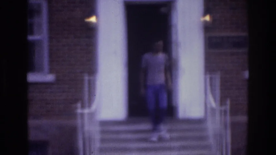 A teenager steps out from a columned brick building in the Atlanta 1975 8mm scene, moving down the exterior stairs.