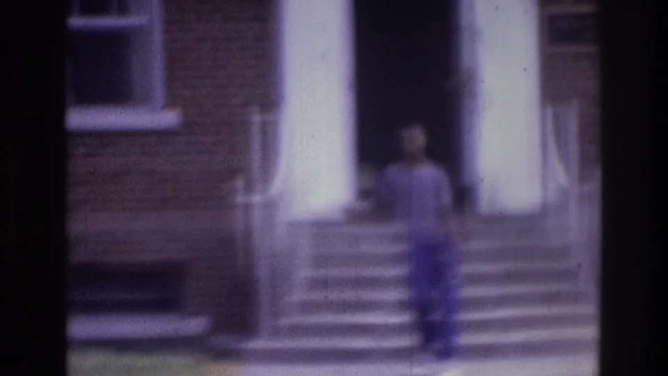 The teenager walks farther from the brick building entrance, with white columns, railings, and steps behind him.