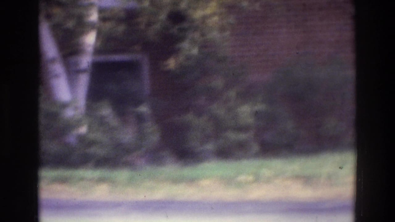 1975 Atlanta home-movie frame showing a grassy residential area, trees, and a brick building in soft focus.