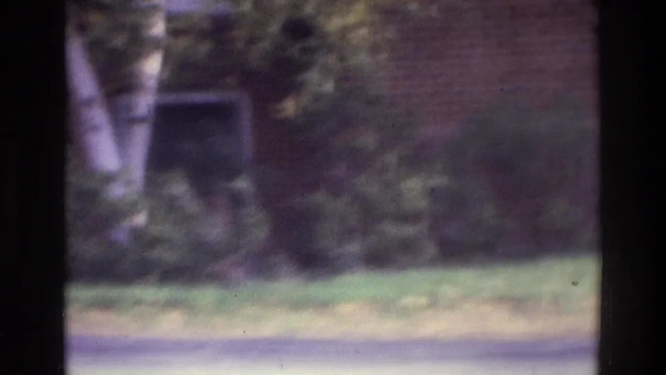 1975 Atlanta home-movie frame showing a grassy residential area, trees, and a brick building in soft focus.