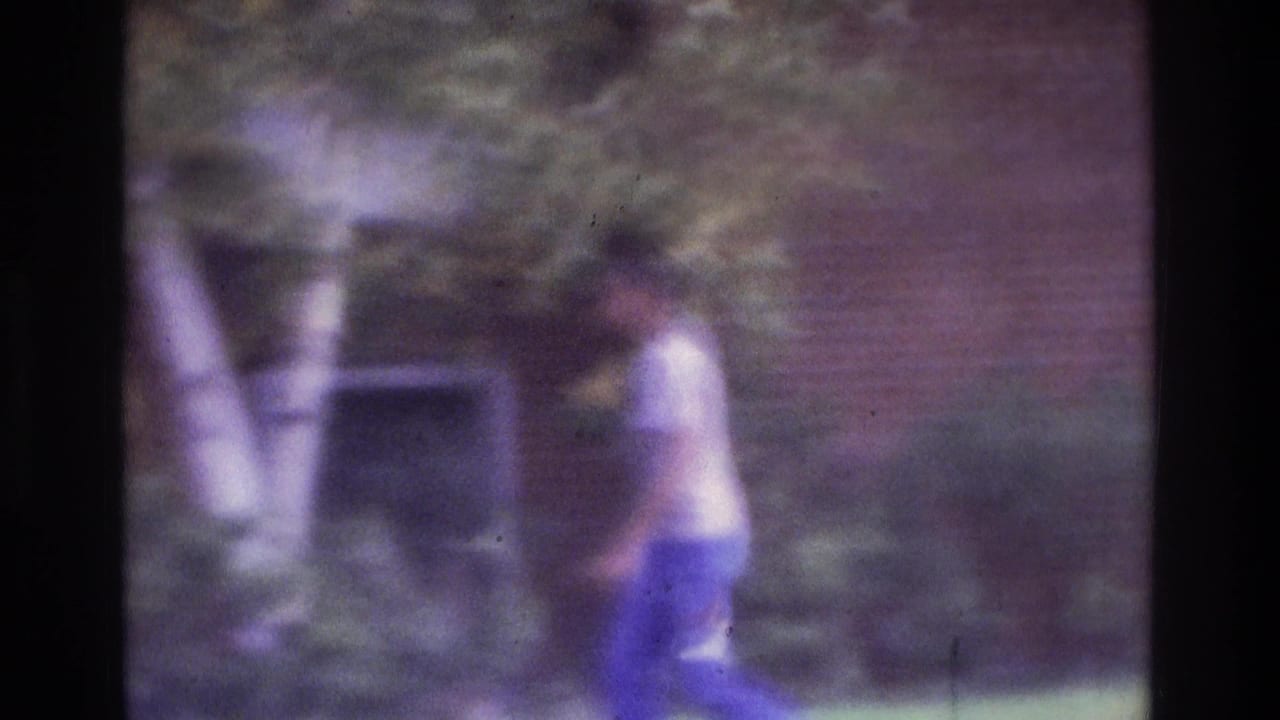 Man in a light shirt and blue pants walking near a brick building and trees.