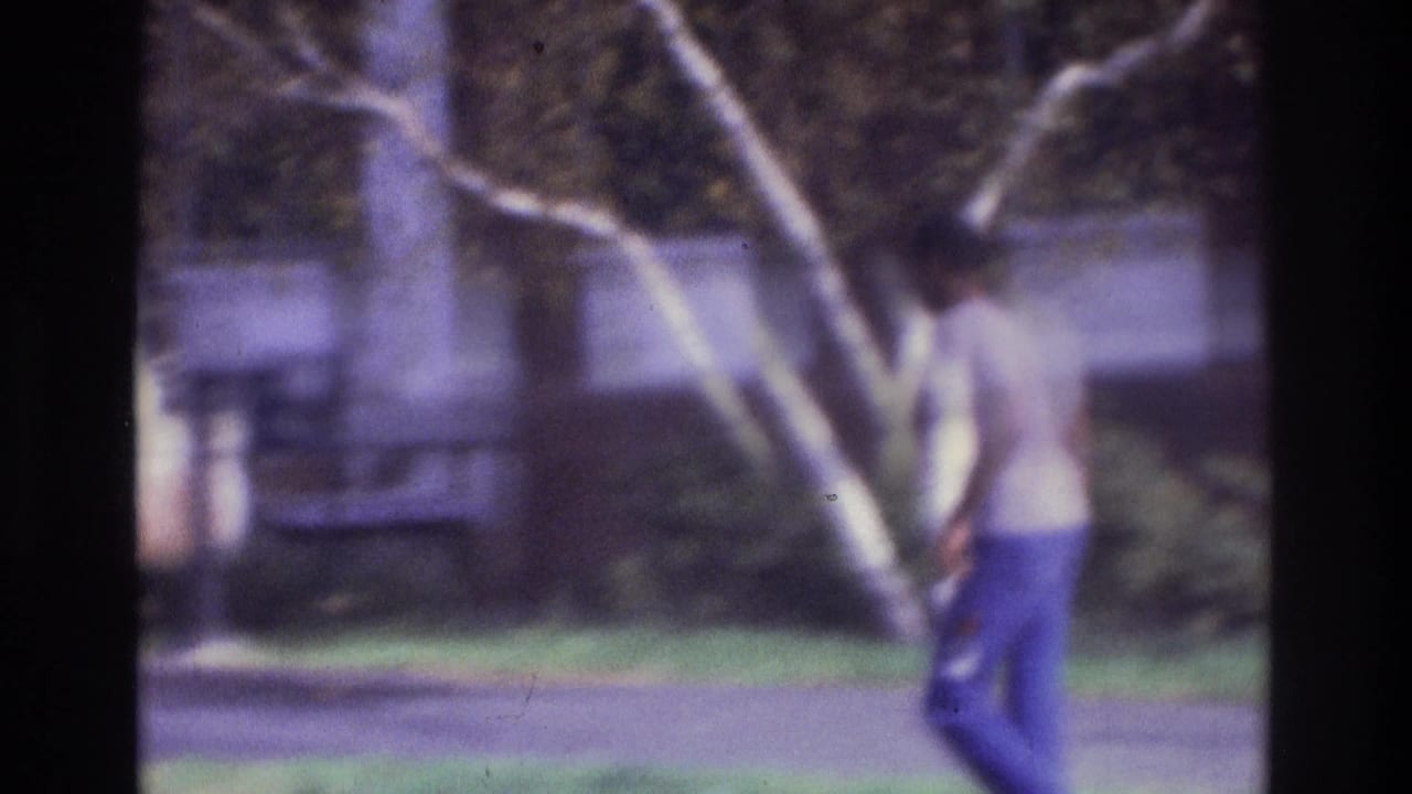 In 1975 Atlanta home-movie footage, a man walks across grass near a white-trunked tree with a house and street behind him.