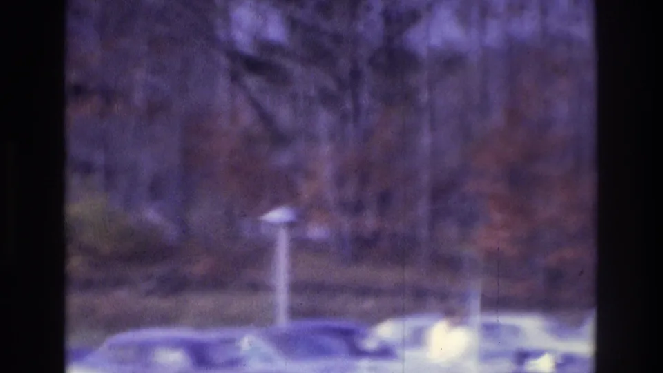 Atlanta 1975 home-movie frame with parked cars near a wooded roadside and a small post or sign in the foreground.
