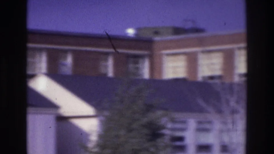 1975 Atlanta 8mm frame with a brick building, roofline, white triangular architectural element, hedge, and soft windows.