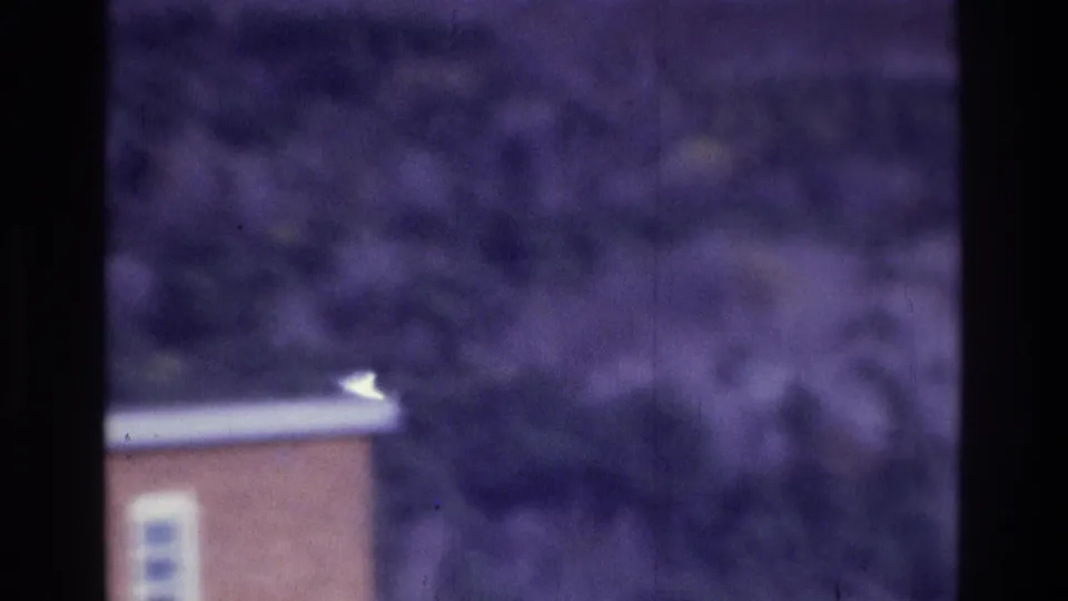 1975 Atlanta 8mm frame showing a low brick building corner, light roof trim, shrubs, trees, and soft purple color shift.