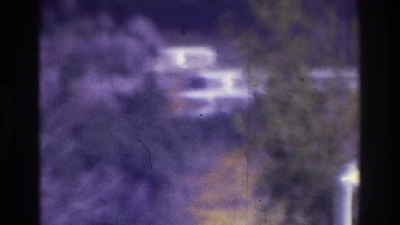Blurred view through a dark vertical frame toward trees, pavement, and indistinct vehicles.