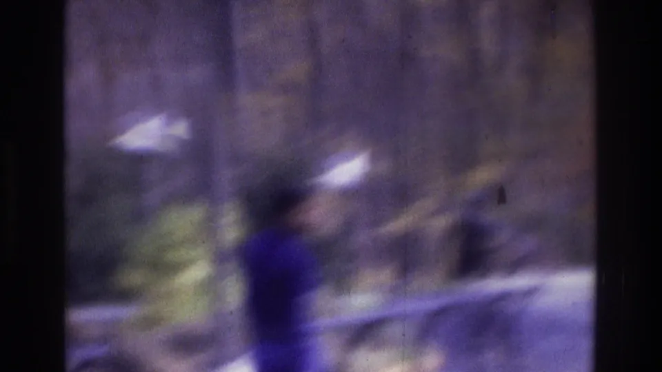 1975 Atlanta 8mm frame showing a blurred walking figure in blue near pavement, trees, rocks, and a roadside guard rail.