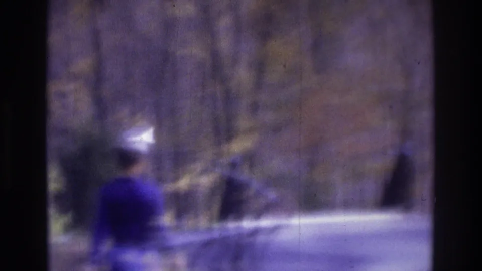 1975 Atlanta home-movie frame with a soft walking figure beside a guard rail in a wooded, rocky roadside setting.